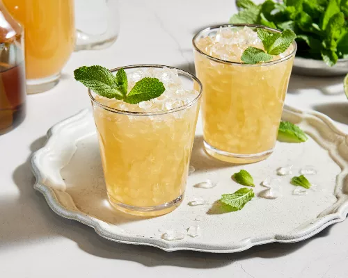 Two glasses of spirit-free mint julep with crushed ice and garnished with mint served on a white platter, shown with a bowl of mint leaves, half a lime, a pitcher of prepared julep and a pitcher of mint simple syrup