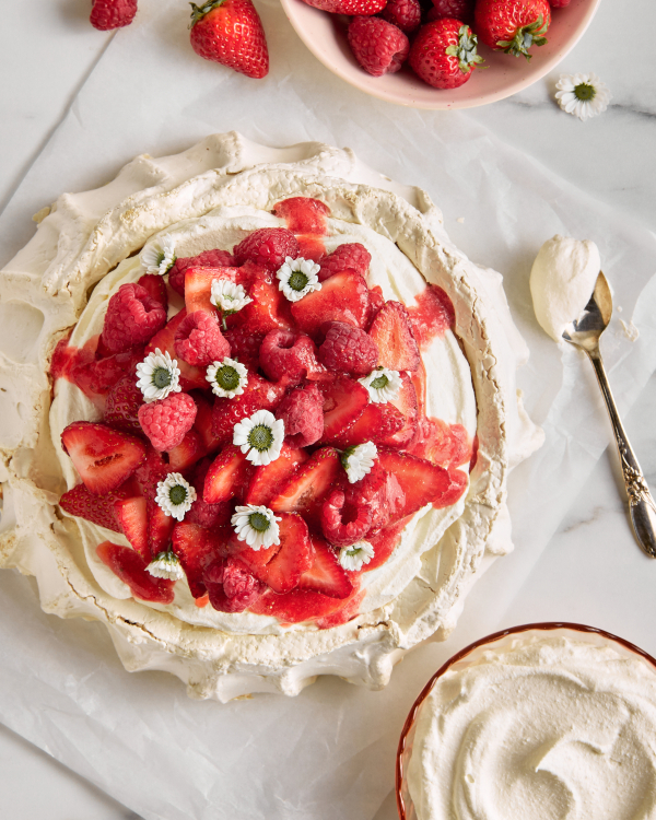 Top view of a Pavlova topped with raspberries and strawberries and flowers, shown with a bowl of berries, a bowl of whipped cream, and a gold spoon