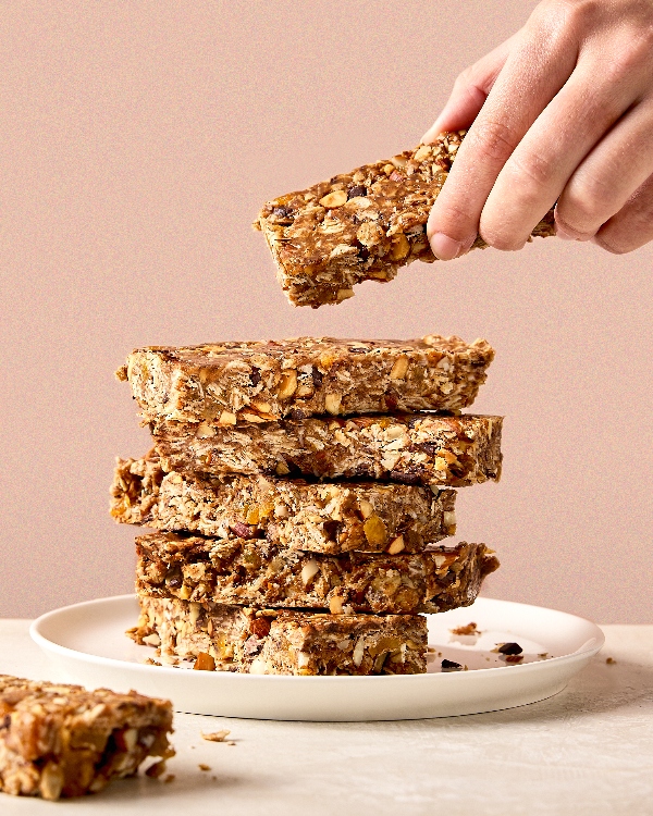 A hand holding a no-bake granola bar above a stack of similar bars on a white plate on a soft pink background. The granola bars, made with oats, nuts, and dried fruits, are rectangular