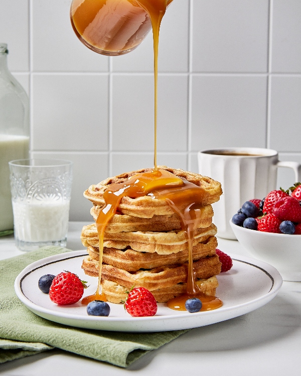 Syrup pouring from a height over a stack of buttermilk waffles on a white plate on a kitchen counter, shown served with a bowl of strawberries and blueberries, a cup of milk, and a mug of coffee.