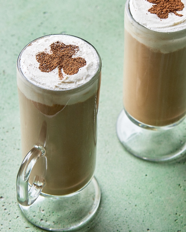 Two Irish whiskey lattes served in tall, clear glass mugs, topped with frothy cream and a clover-shaped sprinkle of cinnamon, perfect for St. Patrick's Day celebrations, set against a textured green background.
