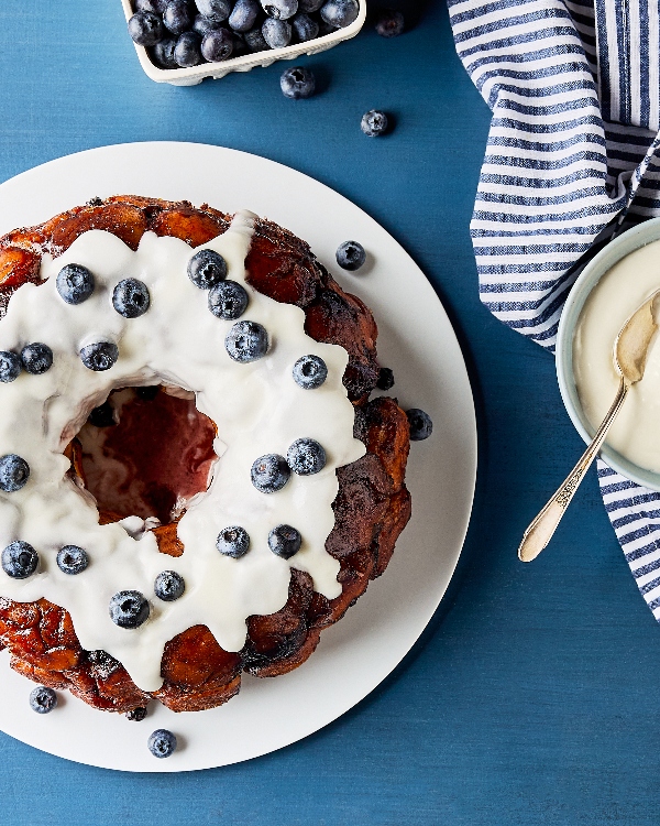 Top-down view of glazed blueberry monkey bread on a white plate, accompanied by a bowl of glaze with a spoon, a basket of blueberries, and a blue-striped napkin, all set on a blue tabletop.