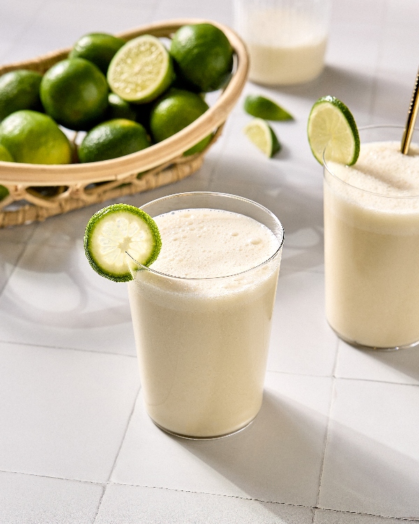 Two glasses of creamy Brazilian limeade, each garnished with a fresh lime slice, sit on a light-coloured tile surface. One glass has a straw, and in the background there is a wicker basket filled with whole limes and lime halves.. Additional lime wedges and a pitcher of limeade are also visible.