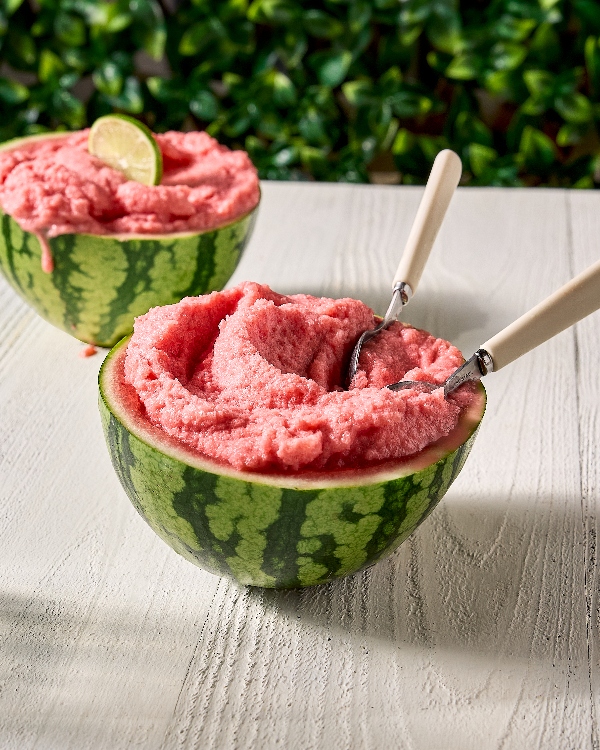 Two hollowed-out watermelon halves filled with smooth, pink watermelon sherbet sit on a white wooden table. The front watermelon half has two resting inside. The back watermelon half is garnished with a lime slice and everything is set against a backdrop of green foliage.