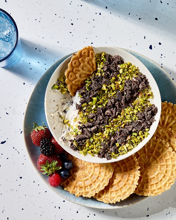 A top-down view of a plate on a limestone counter featuring a bowl of cannoli dip, topped with chopped pistachios and chocolate chunks. Pizzelle cookies and fresh strawberries, blackberries, raspberries, and blueberries, are arranged around the bowl. A glass of sparkling water is visible on the left side.