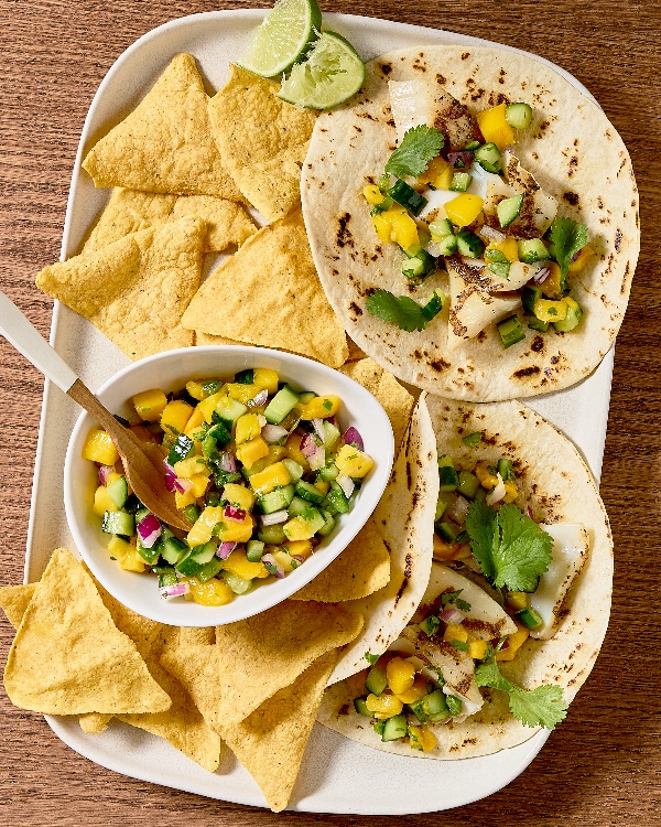 A white plate on a wooden table holds two fish tacos topped with mango cucumber salsa, garnished with fresh cilantro. Next to the tacos, a bowl of salsa with a wooden spoon is surrounded by crispy tortilla chips.