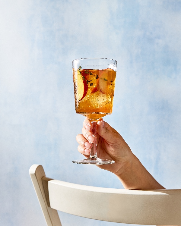 A hand reaching from behind a white patio chair holds up a stemmed glass of peach-thyme sweet tea against a mottled light blue background. The drink is garnished with fresh peach slices and sprigs of thyme, and filled with ice cubes.