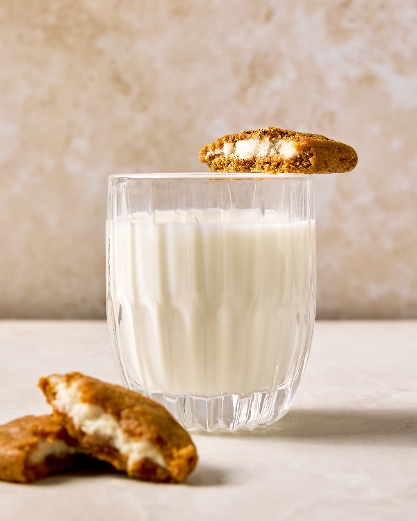 A close-up of a glass of milk with a Pumpkin Spice Cream Cheese Stuffed Cookie resting on the rim. The cookie is broken in half, revealing the cream cheese filling. Another cookie half is placed in front of the glass, highlighting the rich, creamy filling inside the golden-brown cookie