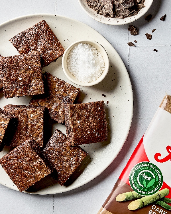 A plate of brownies garnished with flaky salt shown on a white tile counter with a bowl of flaky salt, a bowl of chopped baker’s chocolate, and a bag of dark brown sugar.