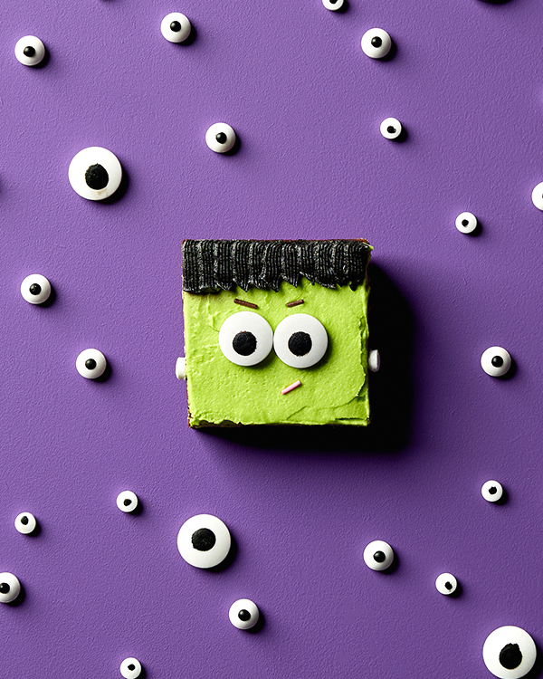 A chocolate brownie decorated with icing and candy eyes to look like Frankenstein’s monster, shown on a purple background with more candy eyes scattered around.