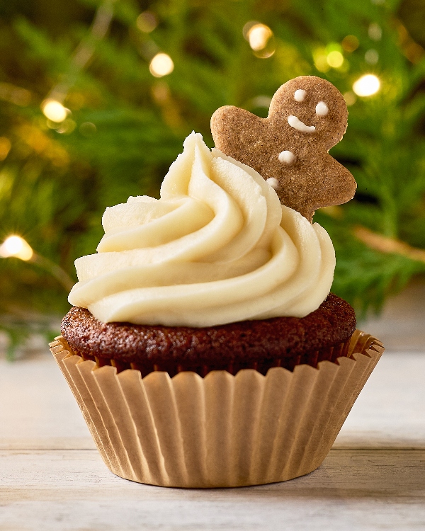 Close-up of a Gingerbread Cupcake with a swirl of white frosting, topped with a mini gingerbread cookie. The background features festive green garland with soft, warm fairy lights, highlighting the holiday theme.