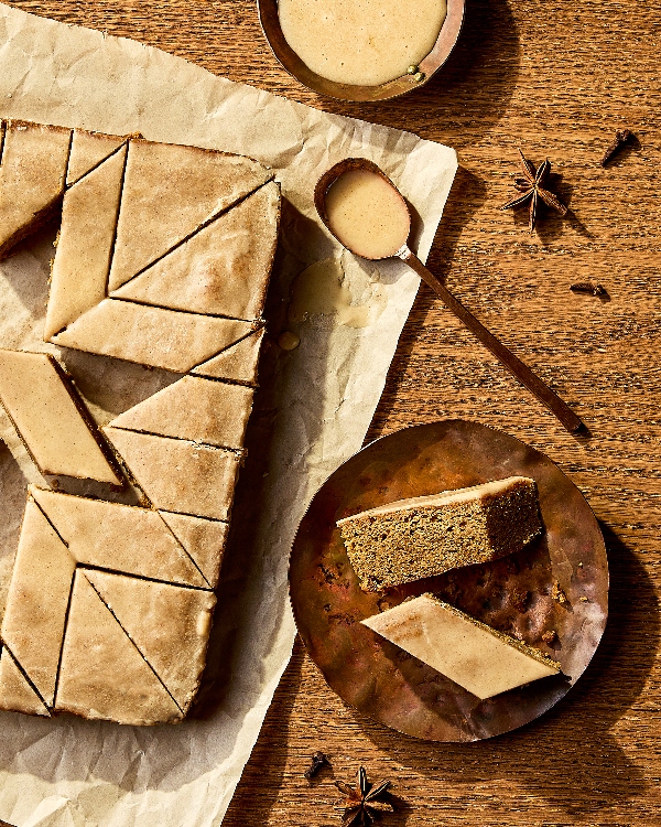 Rows of spiced cake bars cut into diamond shapes, topped with a smooth glaze. One bar is broken in half, revealing a moist and crumbly interior. The warm brown background looks rustic and cozy