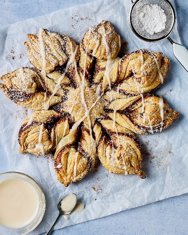 A Glazed Cinnamon Sugar Snowflake pastry displayed on parchment paper, showcasing intricate, twisted layers filled with cinnamon and sugar. The pastry is drizzled with glaze and dusted with powdered sugar, surrounded by a sifter with extra powdered sugar and a bowl of glaze with a spoon on the side.