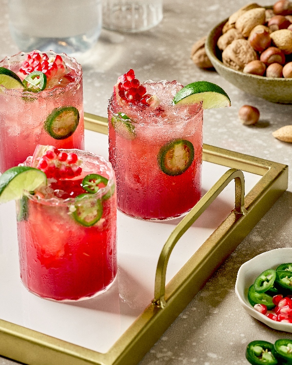 Three glasses of Spicy Pomegranate Ginger Fizz arranged on a gold-rimmed tray, each drink garnished with lime wedges, pomegranate seeds, and slices of fresh jalapeño. The vibrant red cocktails stand out against the tray’s white base, creating an elegant presentation. In the background is a bowl of mixed nuts and a small dish with extra jalapeño slices and pomegranate seeds.