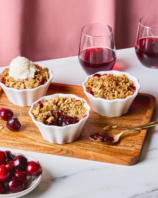 Three servings of Cherry Buckwheat Crisp in scalloped white ramekins, topped with golden oat crumble, one with a scoop of vanilla ice cream. The desserts are set on a wooden tray alongside fresh cherries and two glasses of red wine with a pink background.