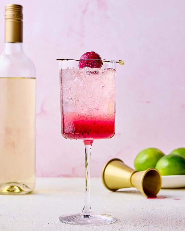 A single glass of dragon fruit spritzer with a pink gradient, served over ice and garnished with a skewered dragon fruit ball. The background features a pale pink hue, a bottle of white wine, a gold jigger, and fresh limes.