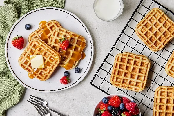  Three buttermilk waffles on a plate served with a pat of butter and strawberries and blueberries, shown on a kitchen counter with more waffles on a wire cooling rack, a glass of milk, and a bowl of mixed berries.
