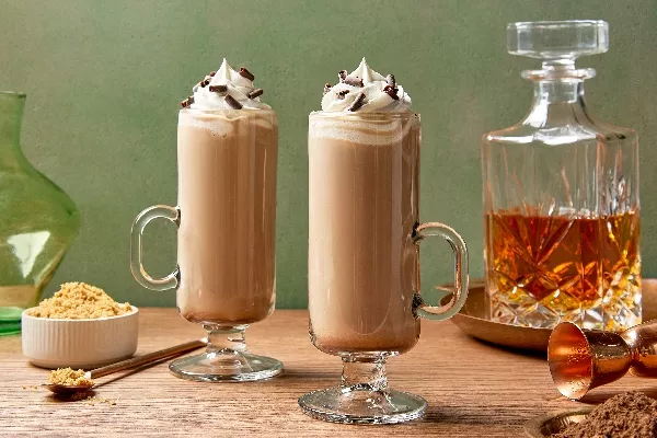 Two tall glasses of Irish whiskey latte topped with whipped cream and chocolate shavings, presented on a wooden table with a side of brown sugar, a decanter of amber whiskey in the background.