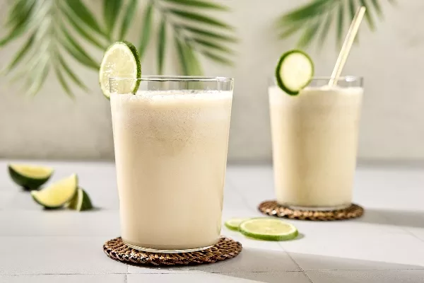  Two glasses of creamy Brazilian limeade are garnished with fresh lime slices and resting on woven coasters. The drinks are set on a light-coloured tile surface, with palm leaves in the background. Additional lime wedges are scattered on the table.