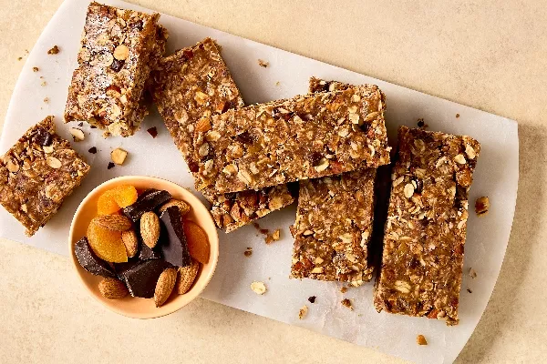 Close-up of homemade no-bake granola bars with oats, nuts, and dried fruits on a white cutting board. A small bowl of almonds, dried apricots, and dark chocolate pieces sits nearby. The granola bars are cut into rectangular pieces.