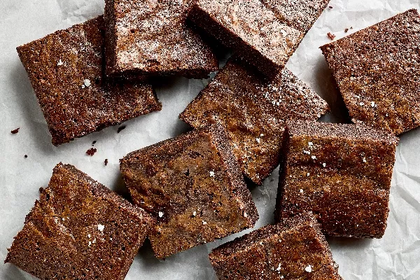 Six square brownies decorated for Halloween like jack o’lanterns, mummies, and Frankenstein, shown on a black plate with bones scattered around