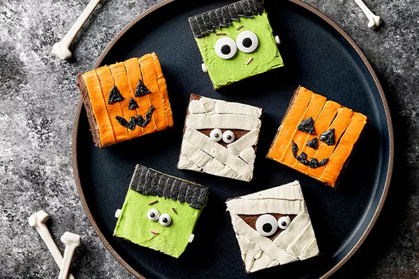 Six square brownies decorated for Halloween like jack o’lanterns, mummies, and Frankenstein, shown on a black plate with bones scattered around.