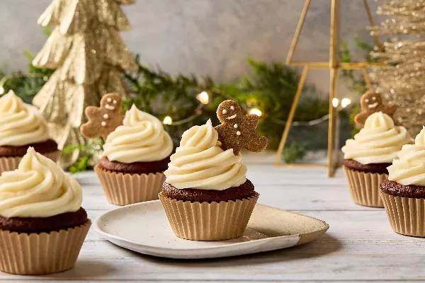 Festive Gingerbread Cupcakes topped with a swirl of creamy white frosting and a mini gingerbread cookie. The cupcakes are arranged on a wooden surface with Christmas decorations in the background, including a gold Christmas tree and twinkling fairy lights.