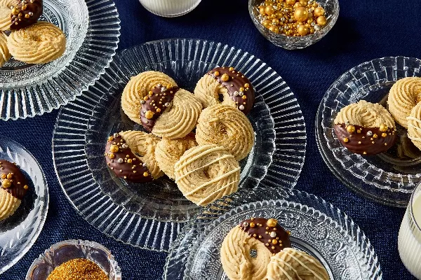 Assortment of piped butter cookies presented on glass plates, showcasing three decoration styles: half-dipped in chocolate with gold sprinkles, drizzled with white chocolate, and sprinkled with golden sugar. The background is a dark blue tablecloth, with additional golden sprinkles in a small dish.