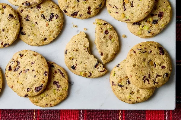 Cranberry pistachio cookies arranged on a white marble surface. The slice-and-bake cookies feature visible chunks of dried cranberries and pistachios, adding texture and colour. One cookie is split in half, showing a tender crumb. A red plaid cloth adds a festive touch at the bottom of the image