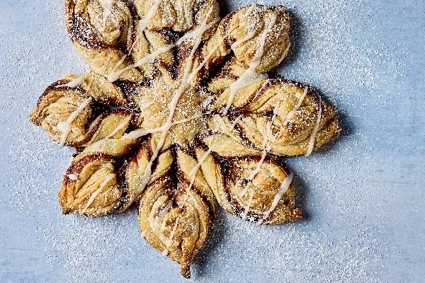 A beautifully crafted Glazed Cinnamon Sugar Snowflake pastry, shaped like a snowflake with twisted layers of dough, filled with cinnamon and sugar. The pastry is drizzled with a light glaze and dusted with powdered sugar, shown on a pale blue background.