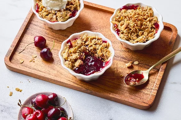 Individual servings of Cherry Buckwheat Crisp in white ramekins, topped with golden oat crumble, one with a scoop of vanilla ice cream. Fresh cherries are scattered on the wooden serving tray, with a bowl of cherries nearby and a gold spoon holding some of the juicy filling, highlighting the dessert's vibrant color and texture.