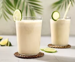  Two glasses of creamy Brazilian limeade are garnished with fresh lime slices and resting on woven coasters. The drinks are set on a light-coloured tile surface, with palm leaves in the background. Additional lime wedges are scattered on the table.