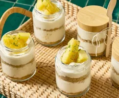  A wicker tray set against a green tiled background holds several jars of single-serving pineapple cheesecake. Each jar features layers of creamy cheesecake and crumbly graham cracker crust, topped with fresh pineapple chunks and a sprinkle of lime zest. Two of the jars have wooden lids, and one has a bamboo spoon tied to it.