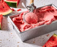 A metal container filled with creamy watermelon sherbet, being scooped with a vintage ice cream scooper. Fresh watermelon slices are scattered around the container on a light-coloured limestone countertop.