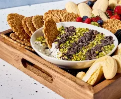 A wooden tray filled with an assortment of Italian cookies and fresh strawberries, blueberries, and blackberries surrounding a bowl of cannoli dip. The dip is topped with chopped pistachios and chocolate chunks and has a Pizzelle cookie dipped in.