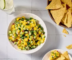 A top-down image of a bowl of mango cucumber salsa on a white tile surface. The salsa also features red onion and cilantro, and a bowl of tortilla chips is placed nearby, with some chips scattered around. A glass of lime-infused water is also visible.