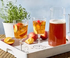 A white tray on a wicker table holds two stemmed glasses, a pitcher of peach-thyme sweet tea, and whole and cut fresh peaches. The drinks are garnished with fresh peach slices and sprigs of thyme, and a potted thyme plant is visible in the background.