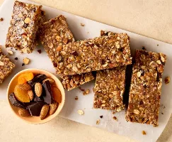 Close-up of homemade no-bake granola bars with oats, nuts, and dried fruits on a white cutting board. A small bowl of almonds, dried apricots, and dark chocolate pieces sits nearby. The granola bars are cut into rectangular pieces.