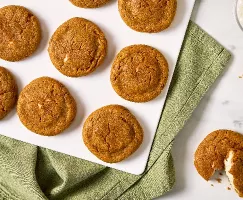 Top view of Pumpkin Spice Cream Cheese Stuffed Cookies arranged on a white tray with a green cloth underneath. The cookies have a golden-brown color and slightly cracked tops, revealing a creamy filling inside. One cookie is broken open, showing the cream cheese filling.